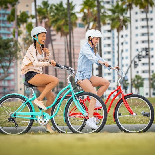 Two women riding bright colored Schwinn cruiser bicycles down a sidewalk on a sunny day.