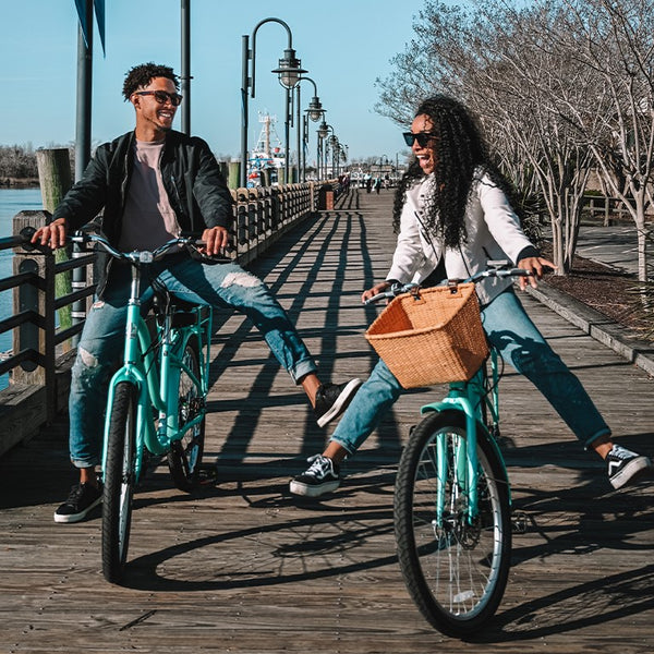 Two people smiling while riding Schwinn bikes down a boardwalk.